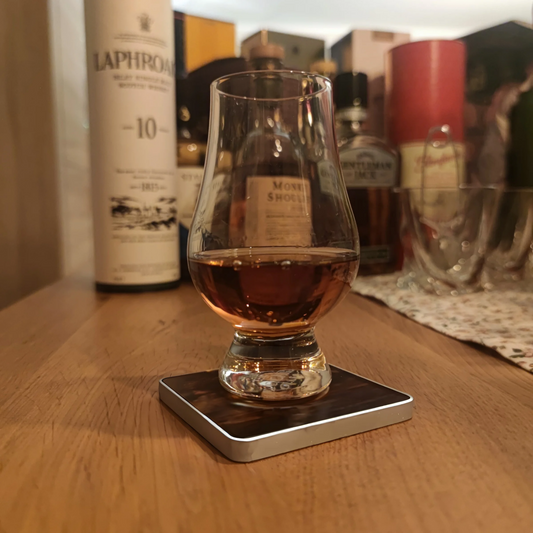 Ein Glencairn Glas mit Whisky auf einem Untersetzer mit Aluminium Rahmen. Im Hintergrund stehen Whiskyflaschen